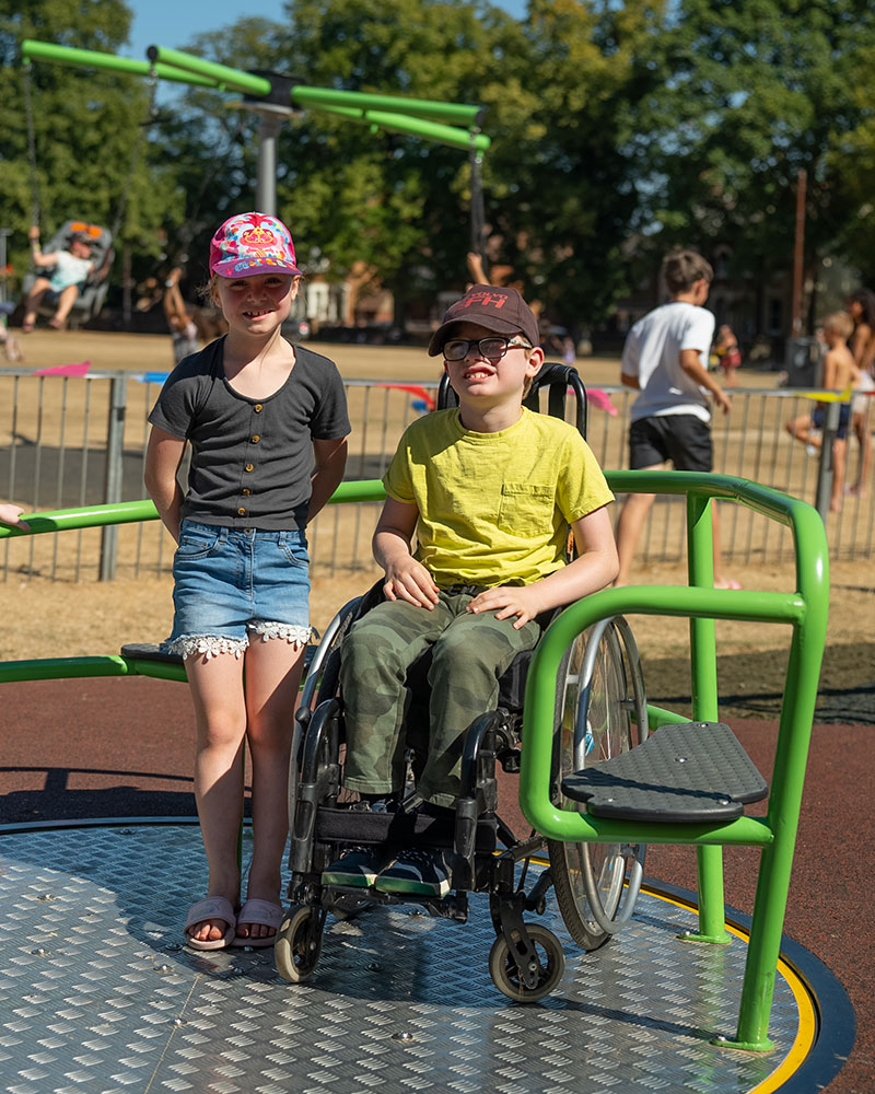 A young boy in a wheelchair, and another young girl are playing together on an inclusive accessible roundabout and smiling at the camera.